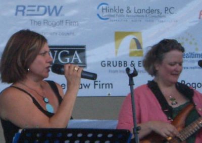 One woman sings with microphone while a woman in the background plays a guitar